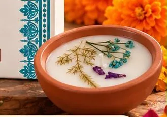 A terracotta wax diya with turquoise buds and fern, set among orange marigolds on a wooden board.